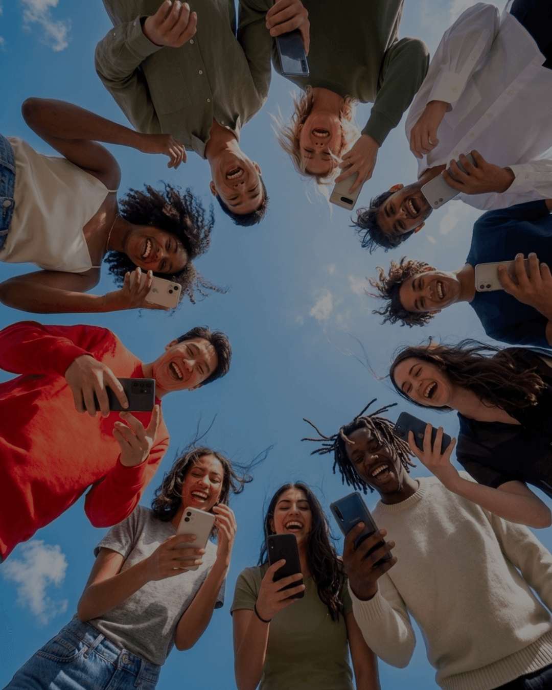 Upward shot of a circle of young people leaning in over the camera against a blue sky, each holding a smartphone and laughing