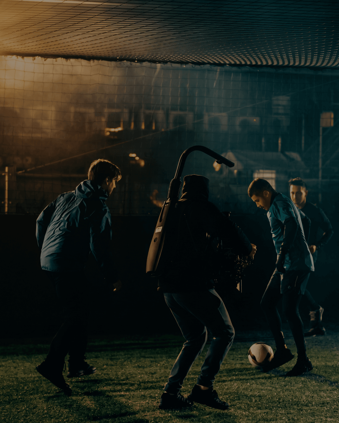 Night shoot on a floodlit five-a-side pitch, a camera operator tracking two players with a gimbal-mounted cinema camera through backlit haze