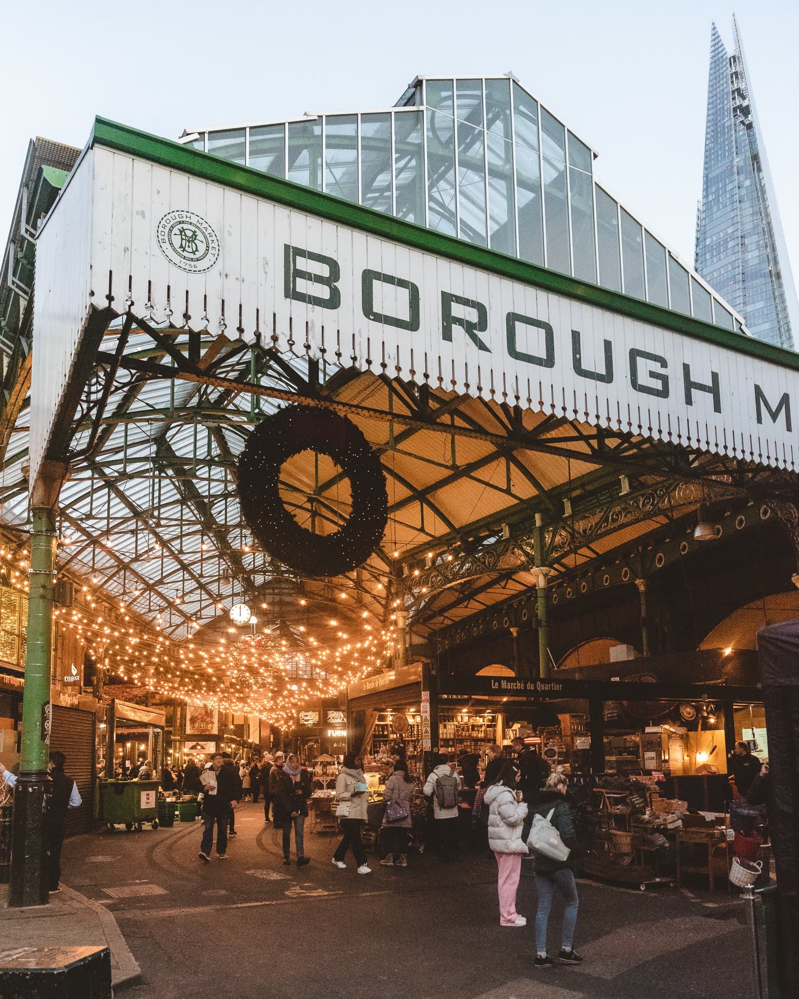 Borough Market's entrance at dusk, a Christmas wreath over the gates, festoon lights, The Shard behind.