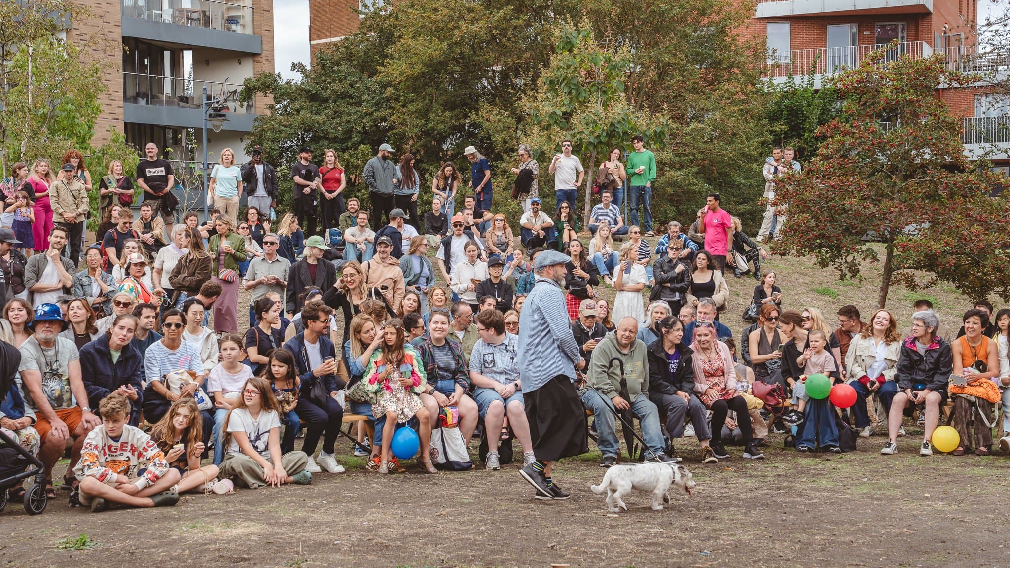 A family audience seated on the grass at the festival, a dog wandering in front.