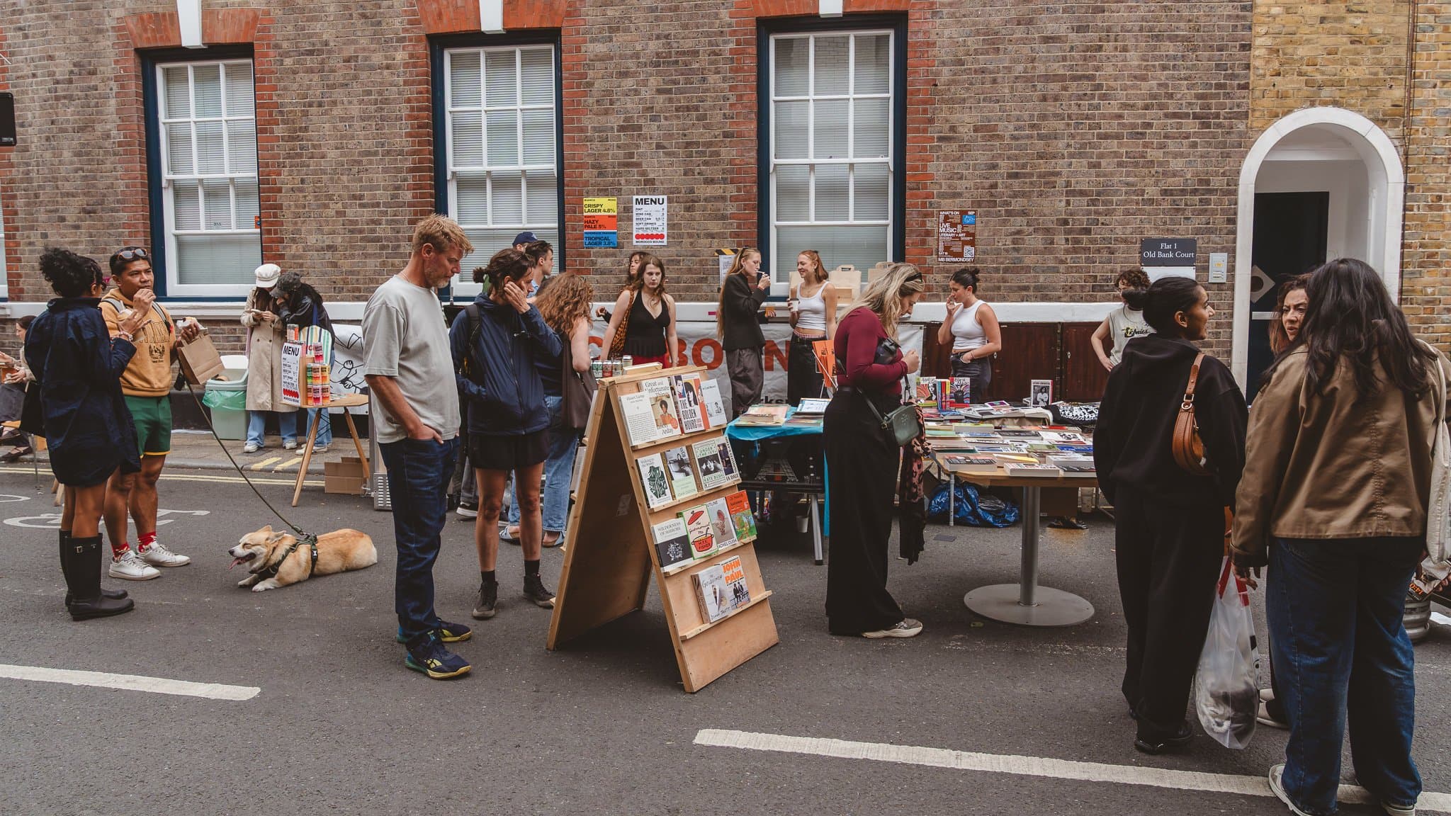 A bookstall on the street, people browsing, a dog at their feet.