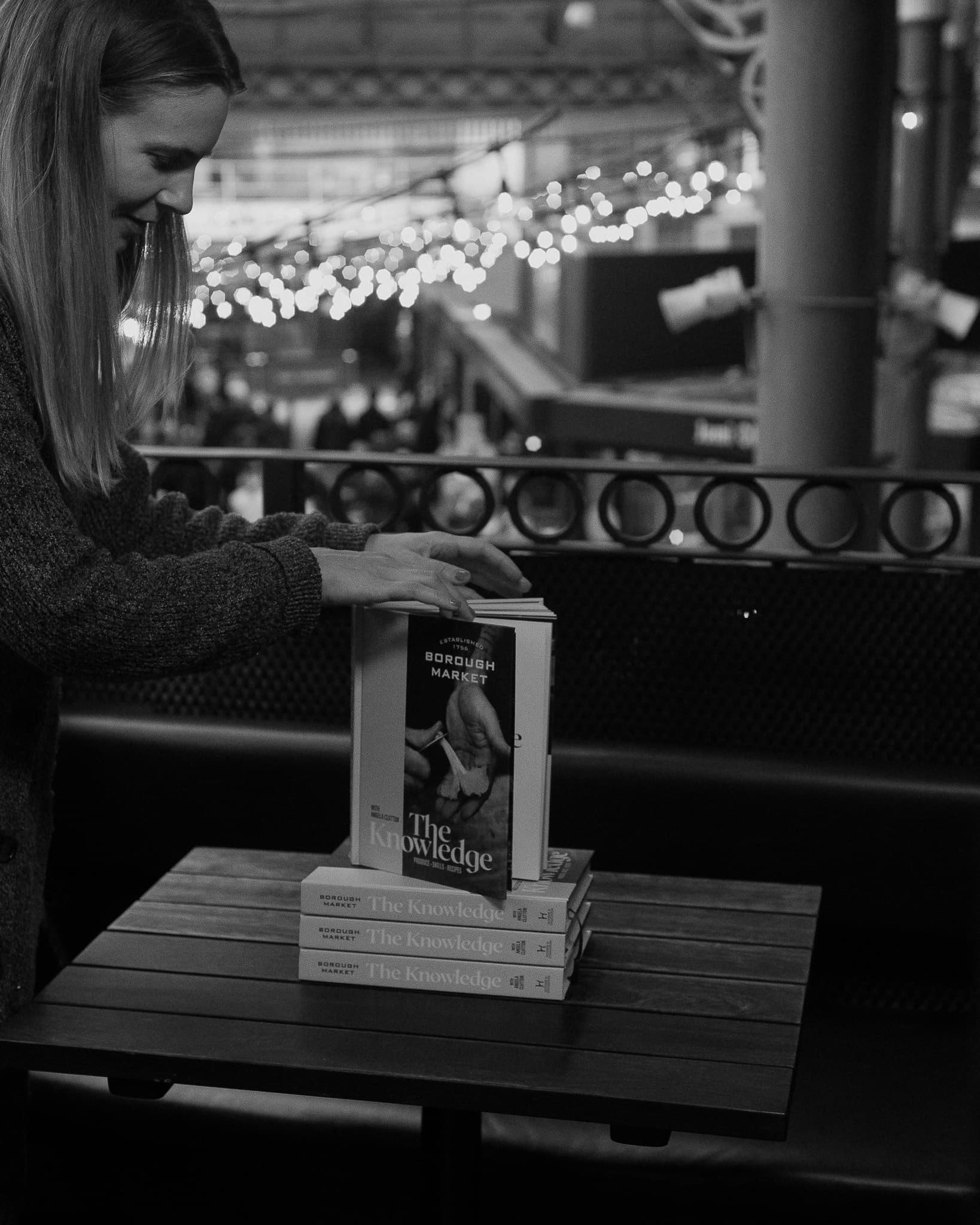 Black and white photograph of a woman stacking copies of the Borough Market cookbook, The Knowledge, on a wooden bench inside the market.