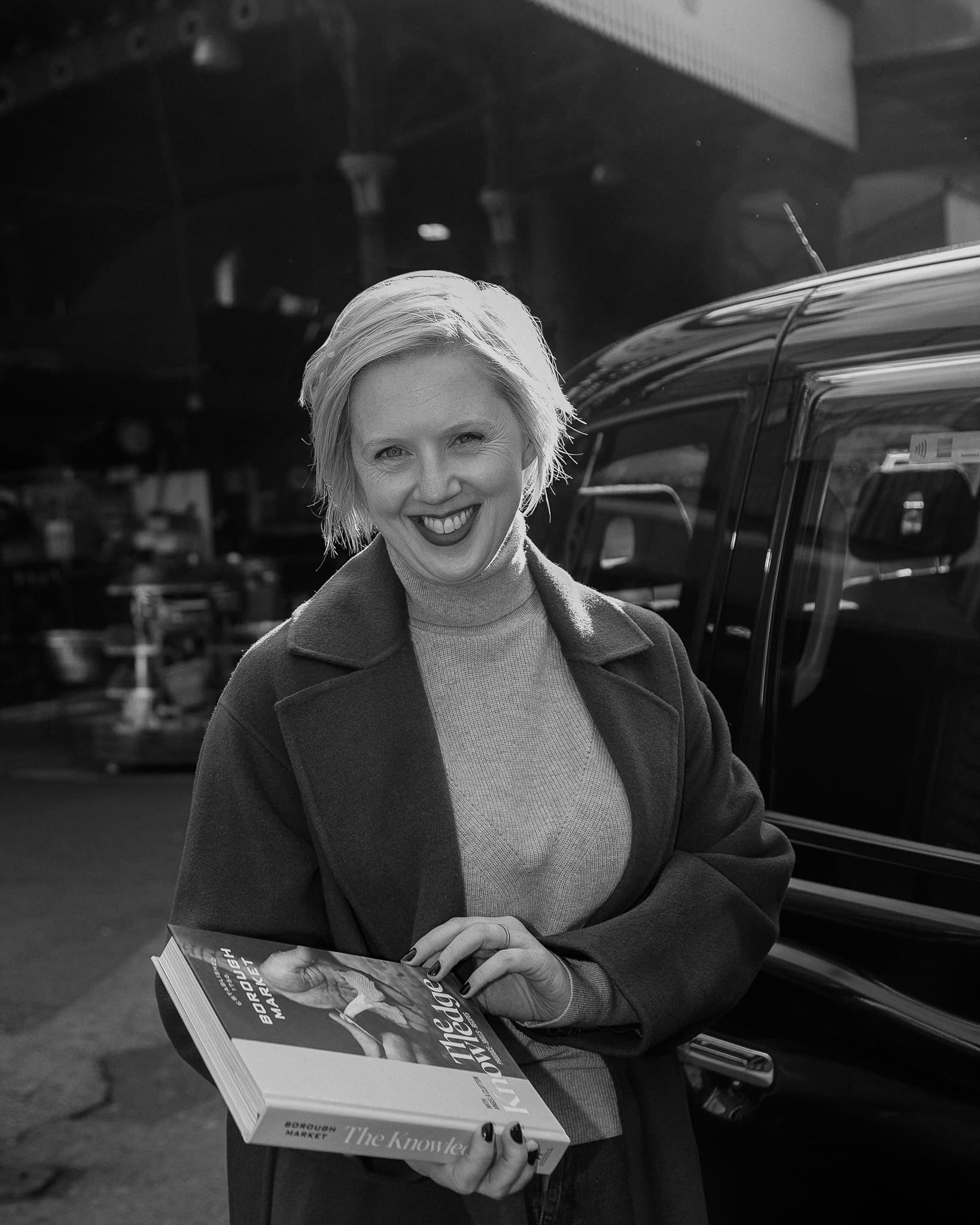 Black and white portrait of a woman smiling and holding a copy of The Knowledge cookbook beside a parked car.