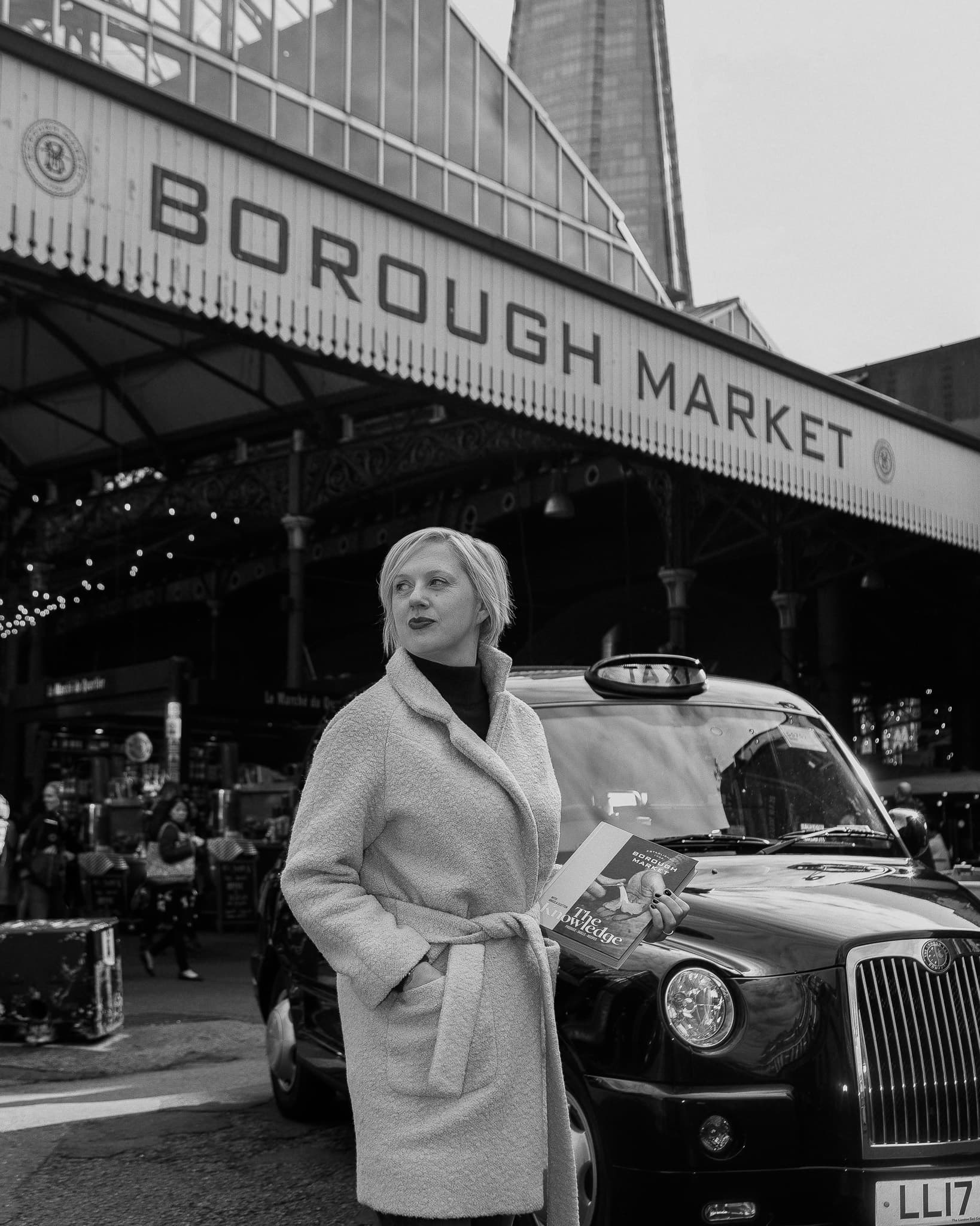 Black and white photograph of a woman holding The Knowledge cookbook in front of the Borough Market entrance sign, the Shard behind her.