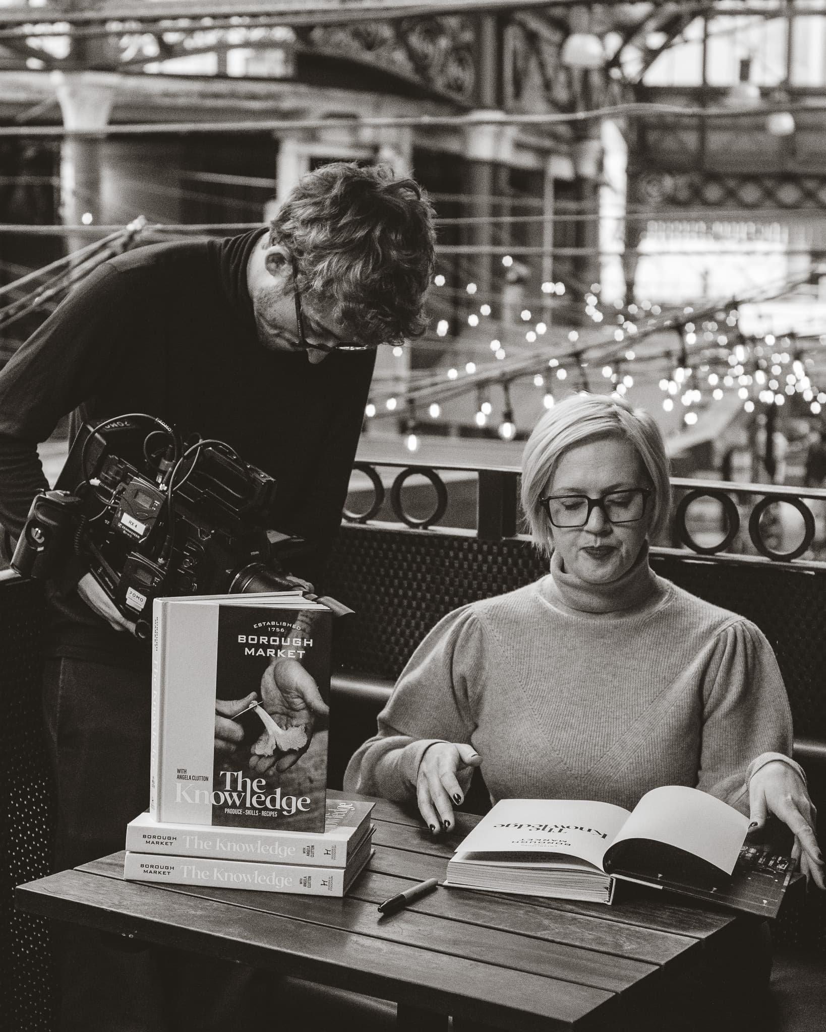 Black and white photograph of a film crew recording a woman reading The Knowledge cookbook at a market table.
