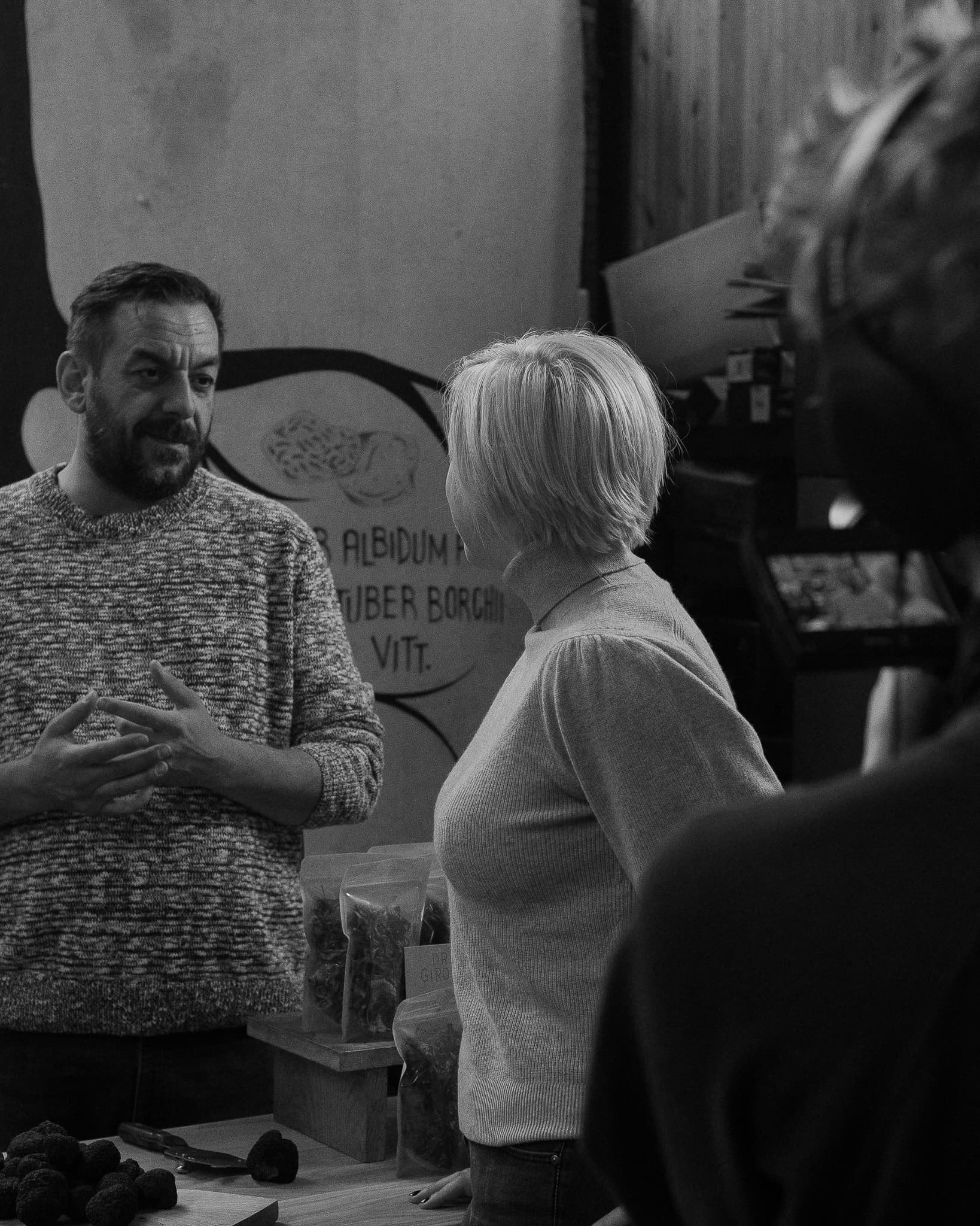 Black and white photograph of a trader talking with a woman inside a specialist truffle shop, hand-painted truffle-species signage and packets of dried mushrooms around them, filmed for the campaign.