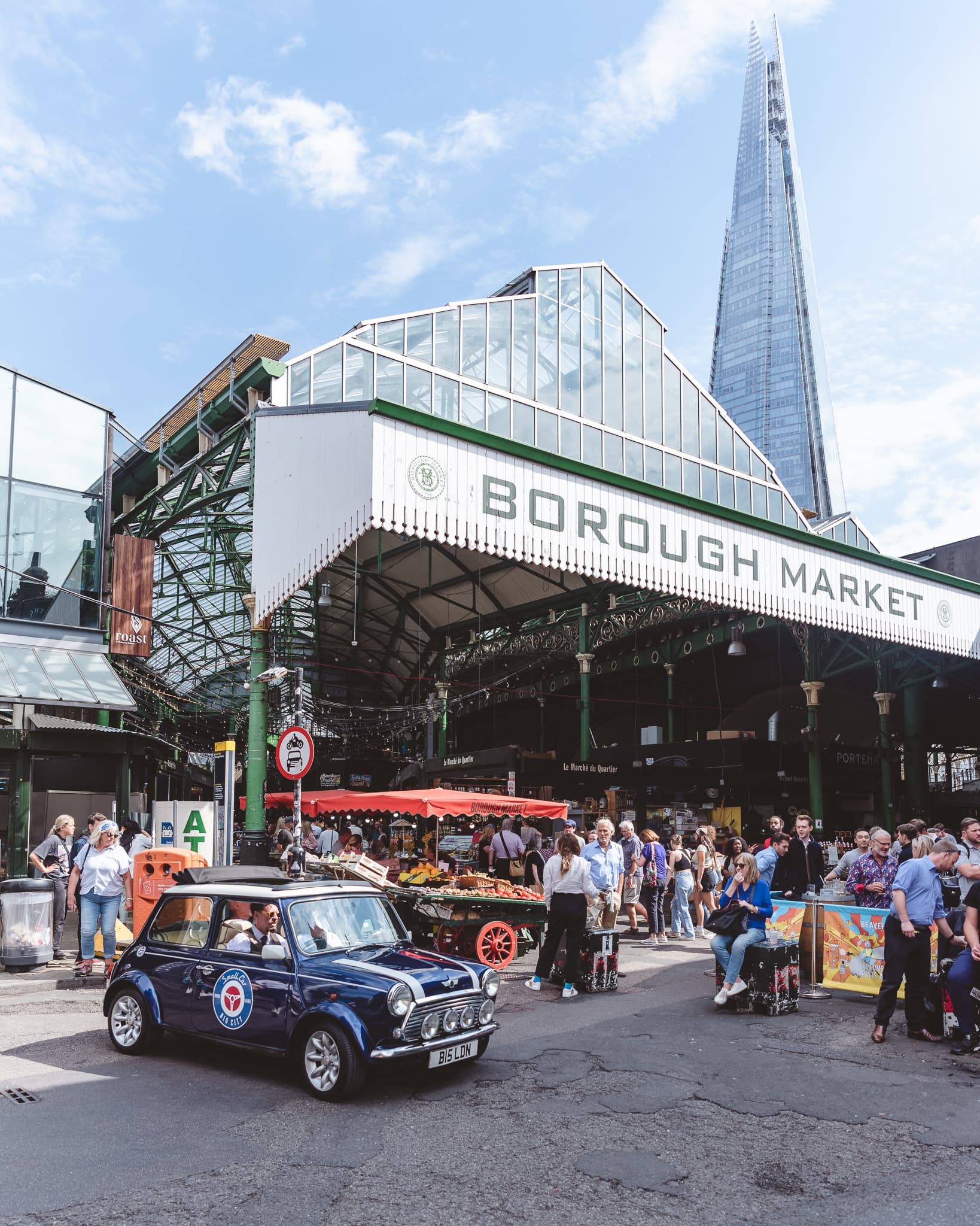 Borough Market's main entrance beneath the railway arches, with the Shard rising behind it and a vintage Mini Cooper parked out front.