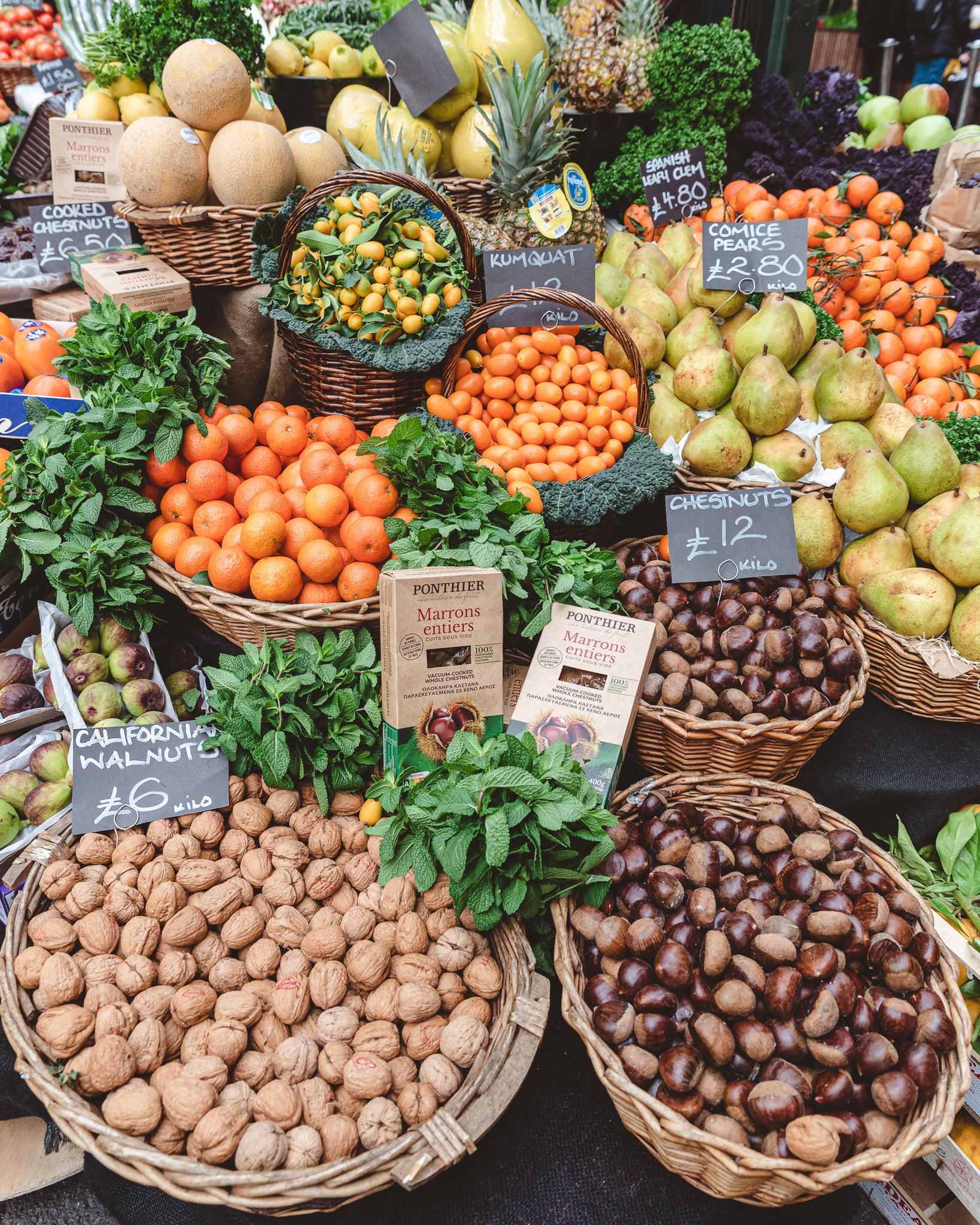 A fruit stall piled with melons, kumquats, satsumas, comice pears, chestnuts and walnuts, each marked with a hand-written price card.