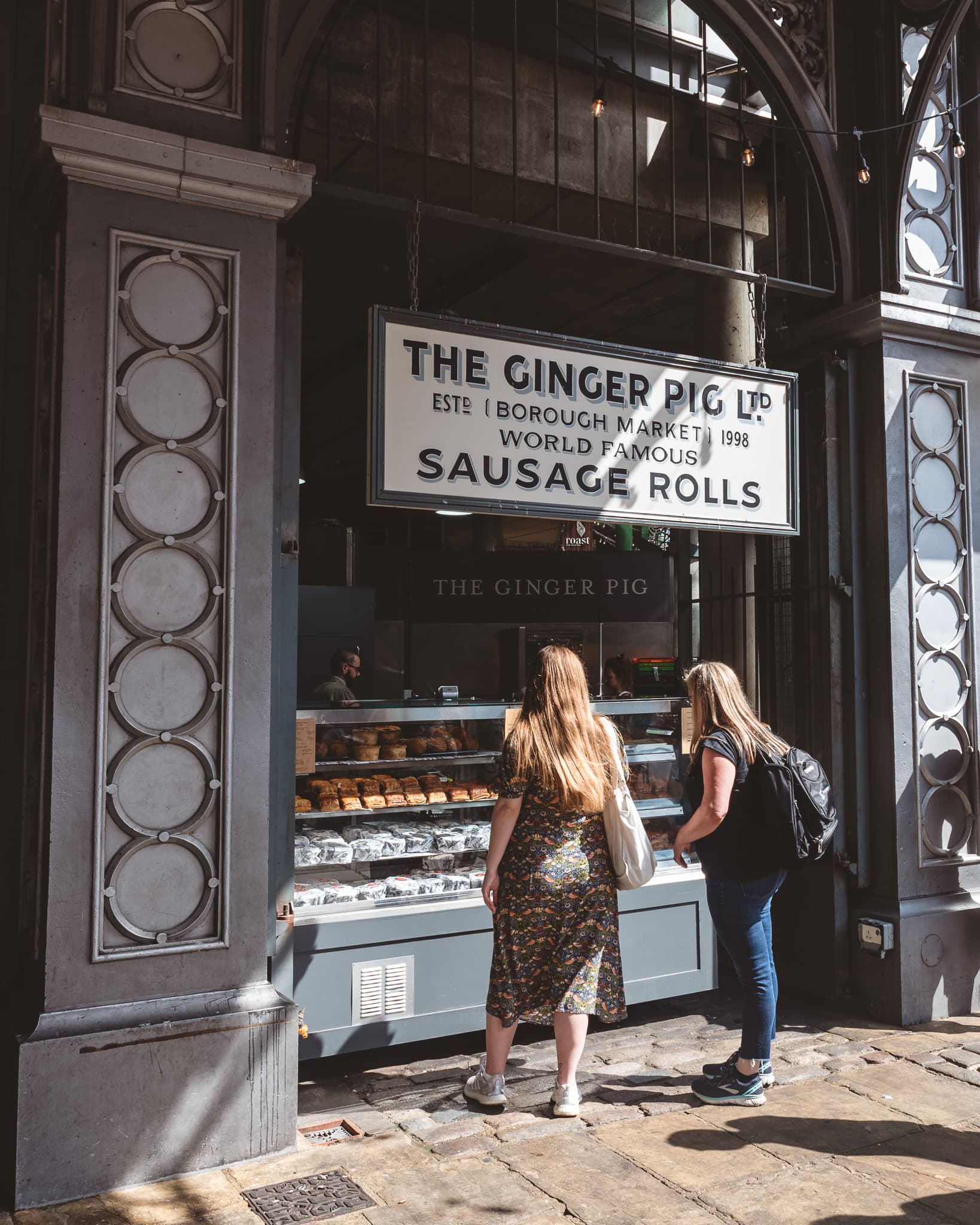 The Ginger Pig's sausage roll stall sign, with two shoppers browsing the pastry case beneath it.