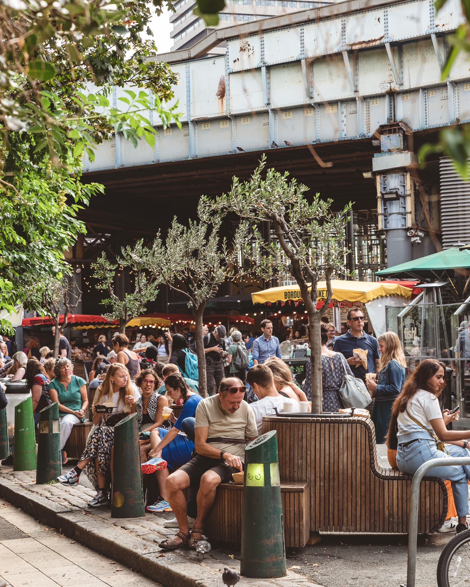 Shoppers seated at outdoor benches under the railway bridge, among olive trees and market stall awnings.