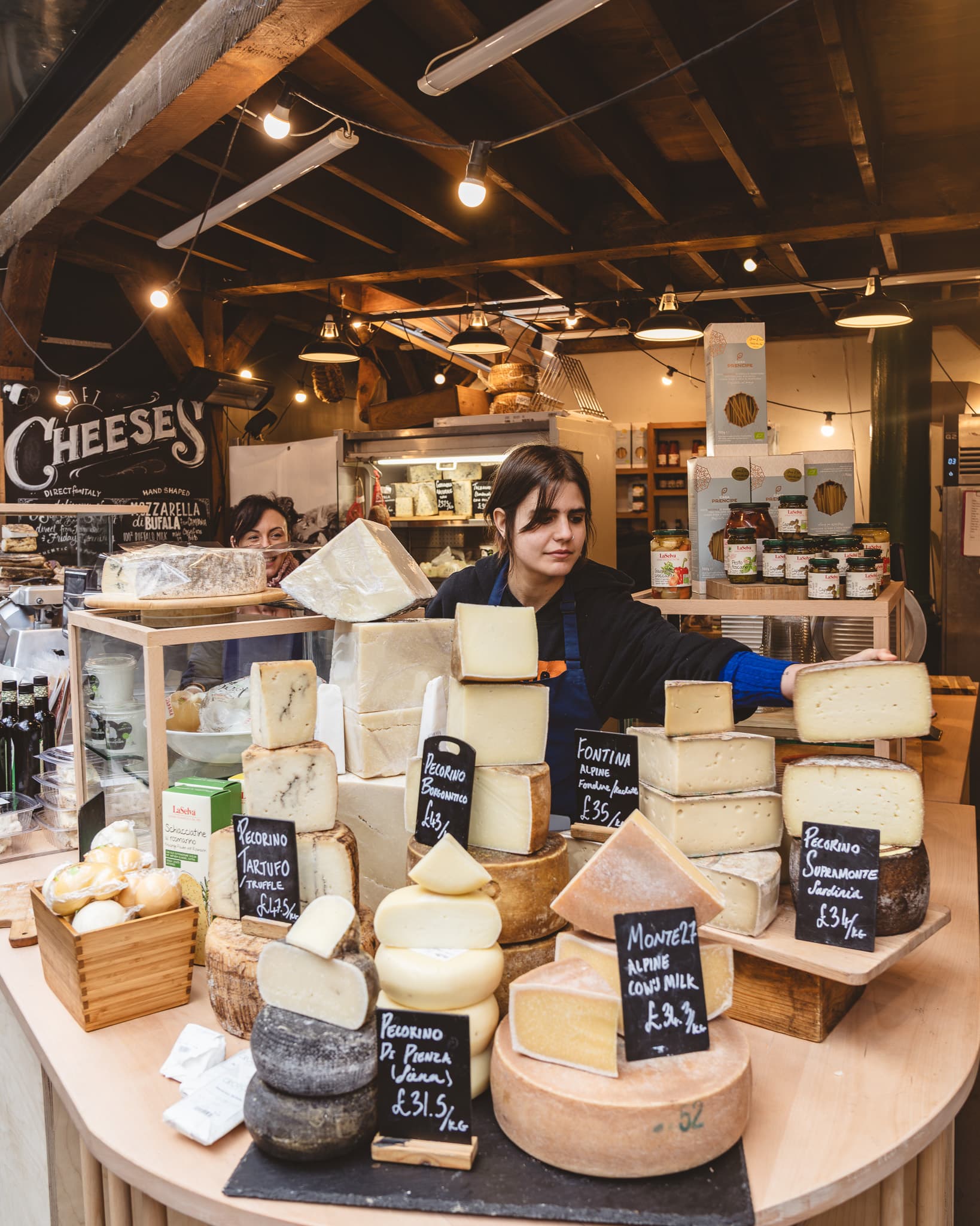 A cheese trader arranging wedges of pecorino and fontina on a market stall, price cards propped among the wheels.