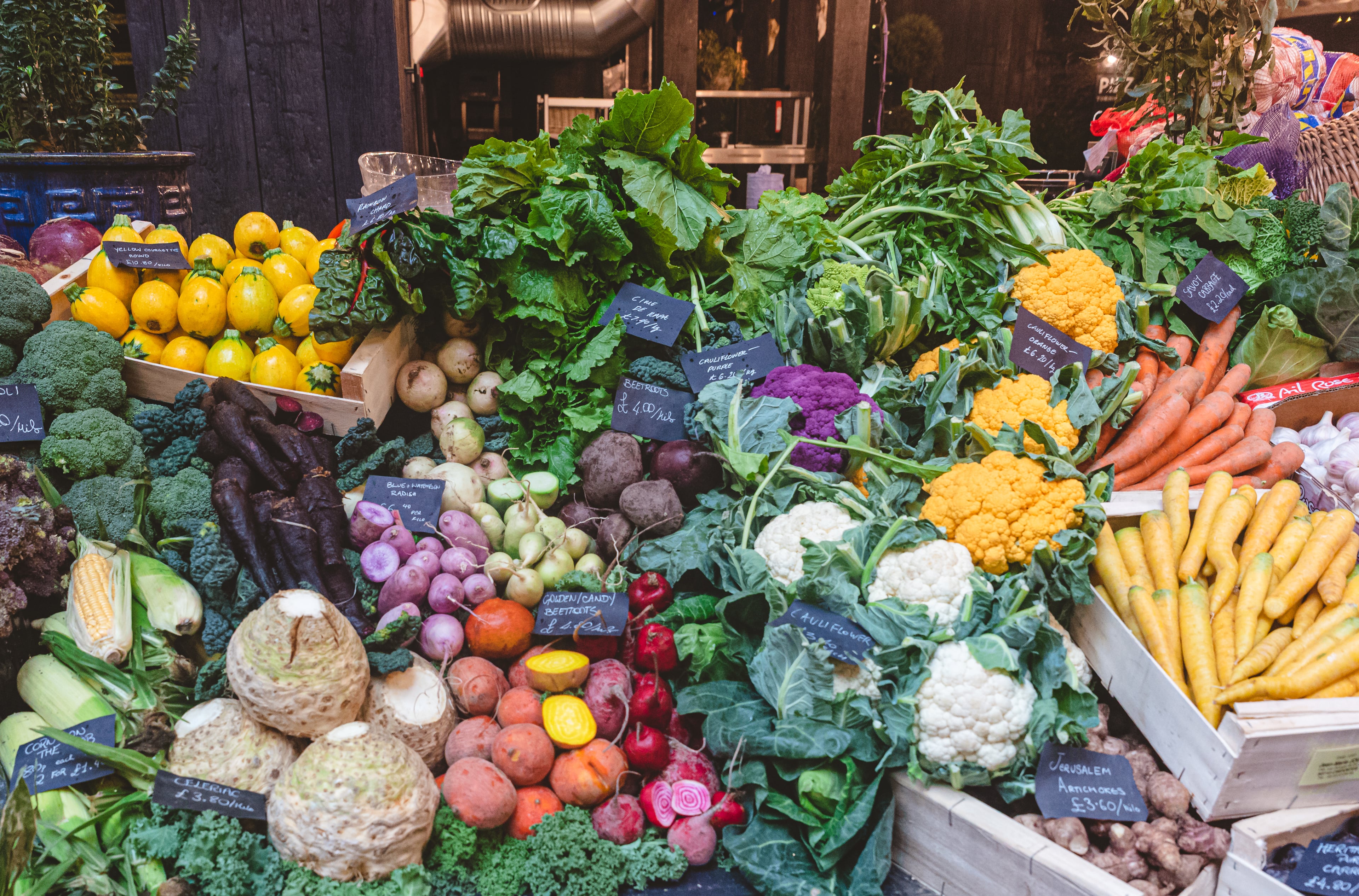 A colourful vegetable stall at Borough Market, with yellow courgettes, purple cauliflower, rainbow chard, carrots and beetroot laid out beneath hand-written price cards.
