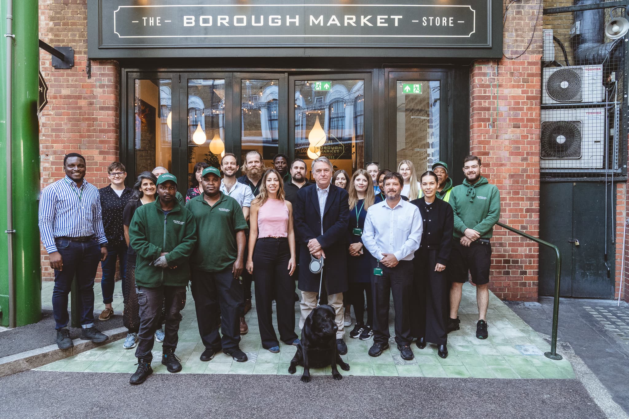 Borough Market's staff team posing outside The Borough Market Store entrance, with a black Labrador in the foreground.