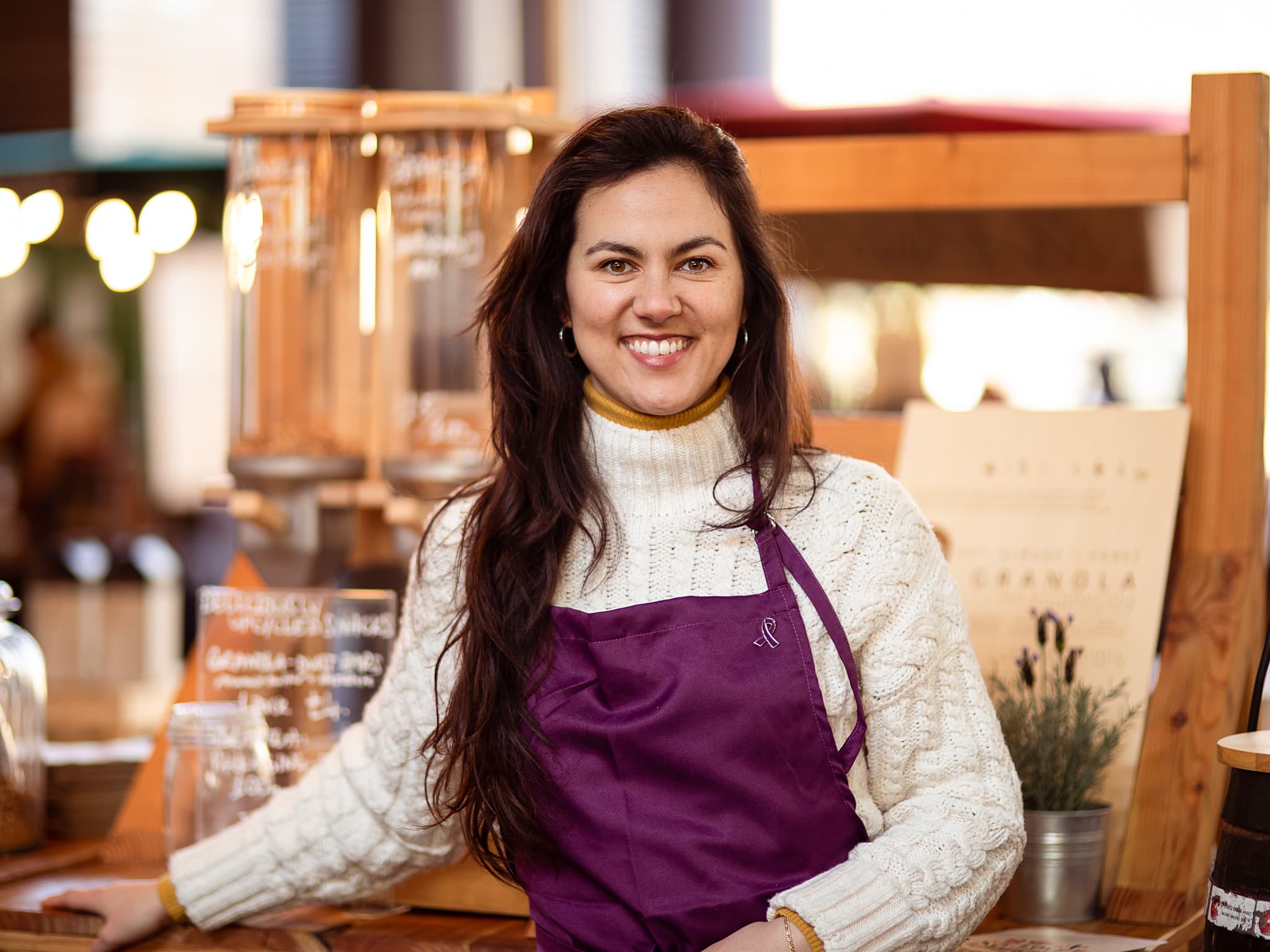 A trader in a purple apron smiling behind a stall selling granola and oat-based products.