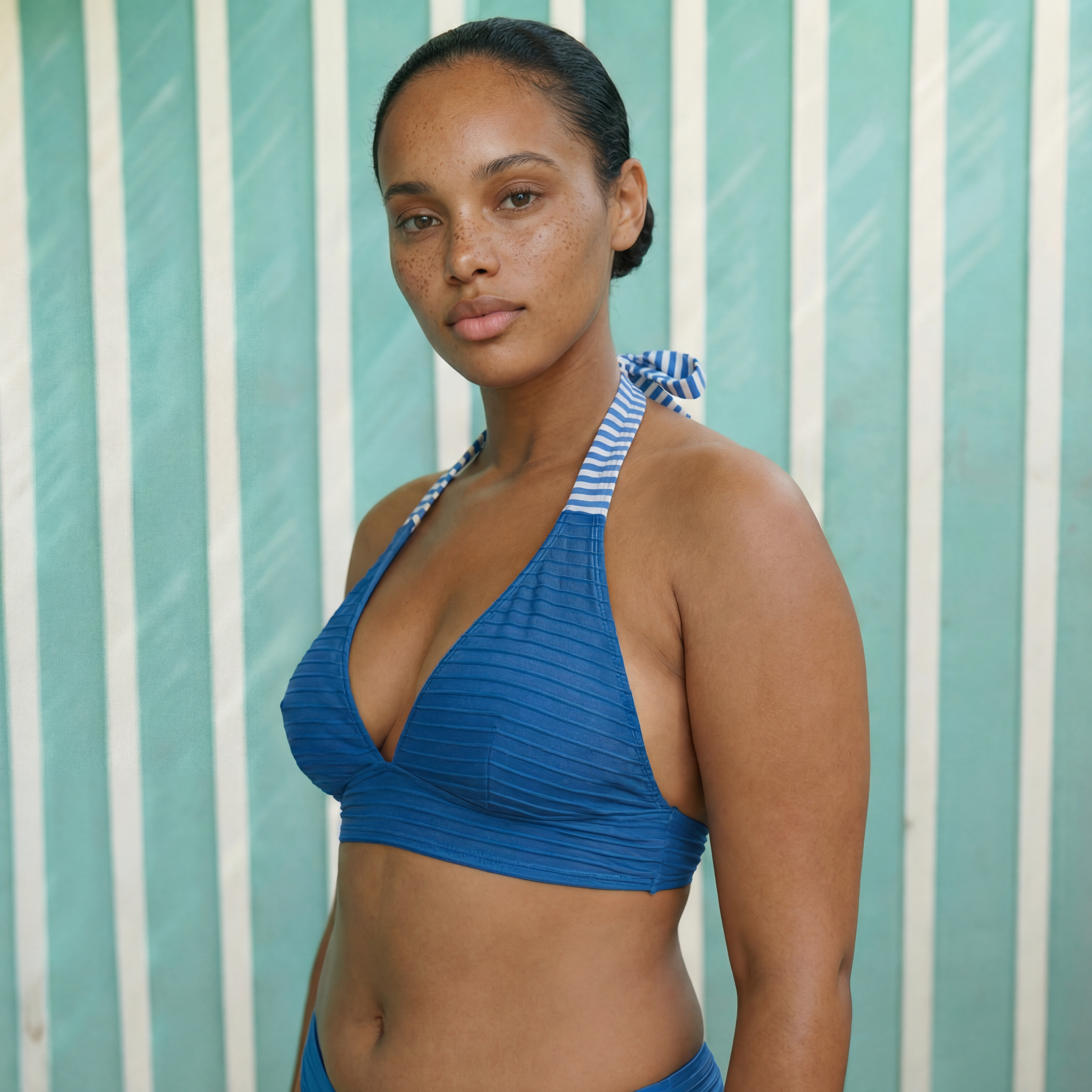 Ecommerce frame: model in a blue ribbed halter bikini top, turned three-quarters to camera against striped fabric