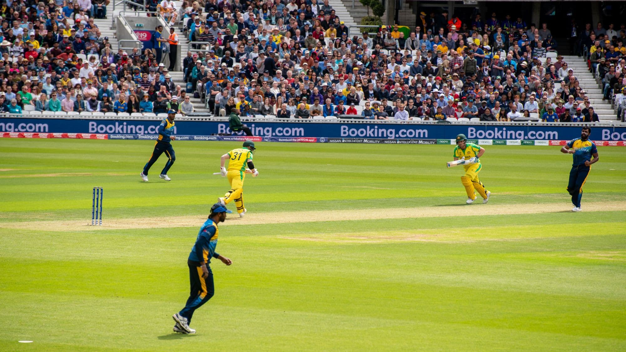 Fielders and batters mid-play at a packed cricket ground, boundary boards lining the pitch.