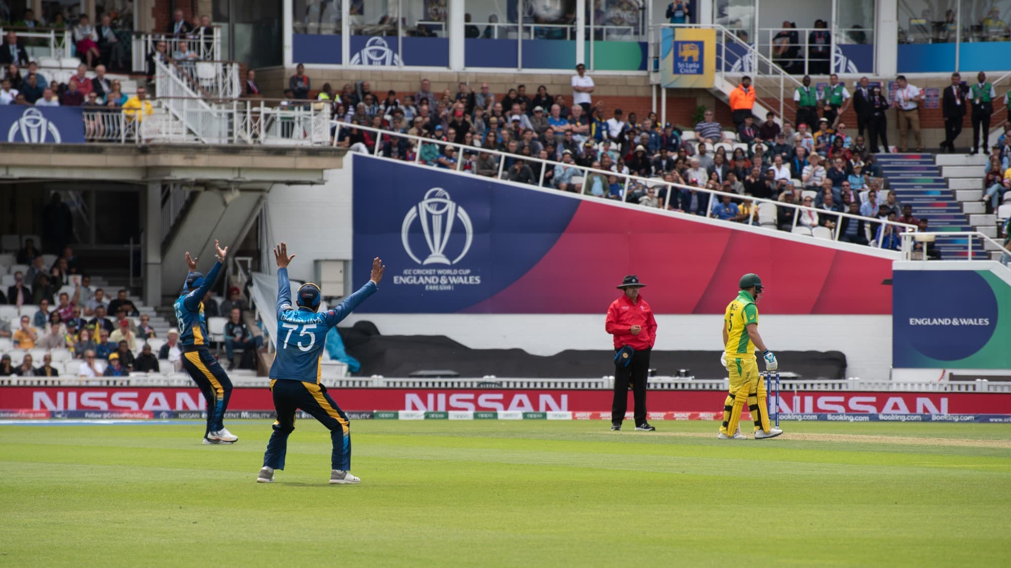 Fielders appeal for a wicket as a batter walks off, Cricket World Cup boards behind them.
