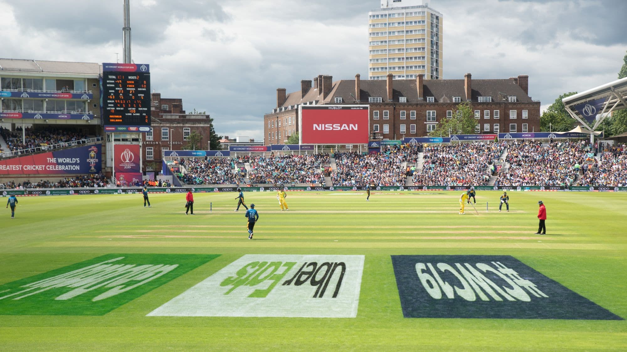 A wide view of a cricket ground mid-over, with Uber Eats branding painted pitch-side on the outfield.